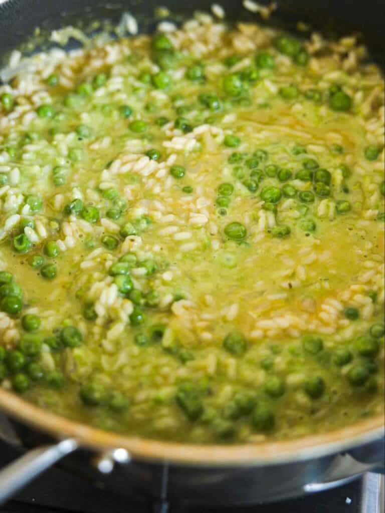 A close-up of a pan filled with creamy risotto, featuring green peas and rice simmering in a yellowish broth.
